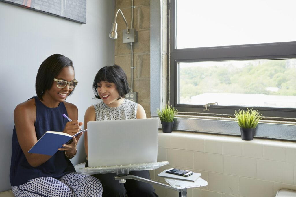 Woman working on laptop, on laptop stand, colleague sitting beside her writing in notebook